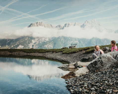 Kinder sitzen am steinigen Ufer eines  Sees mit bergiger Landschaft im Hintergrund 