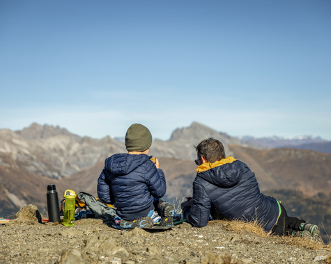 Zwei Wanderer sitzen auf einem Aussichtspunkt und blicken auf die umliegenden Berge.