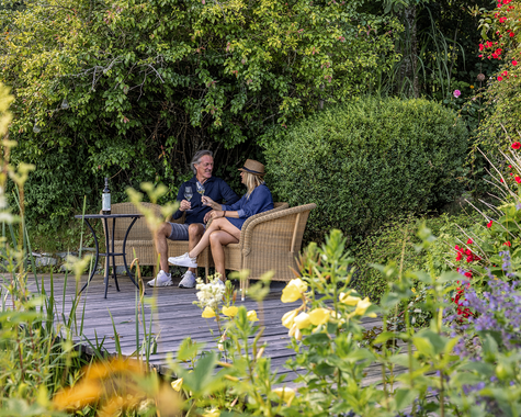 Zwei Personen sitzen an einem Tisch in einem Garten mit Blumen und Sträuchern.