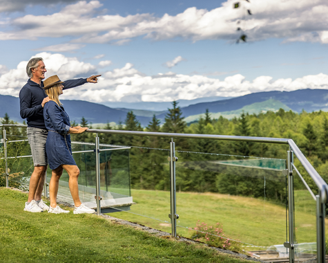 Zwei Personen stehen an einem Geländer mit Blick auf eine Landschaft und Berge im Hintergrund.