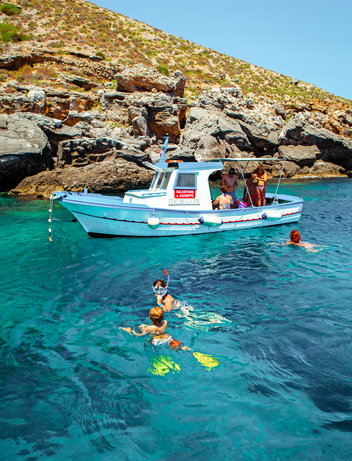 Kleines Boot ankert in einer geschützten Bucht, während Gäste im klaren Wasser schwimmen und schnorcheln.