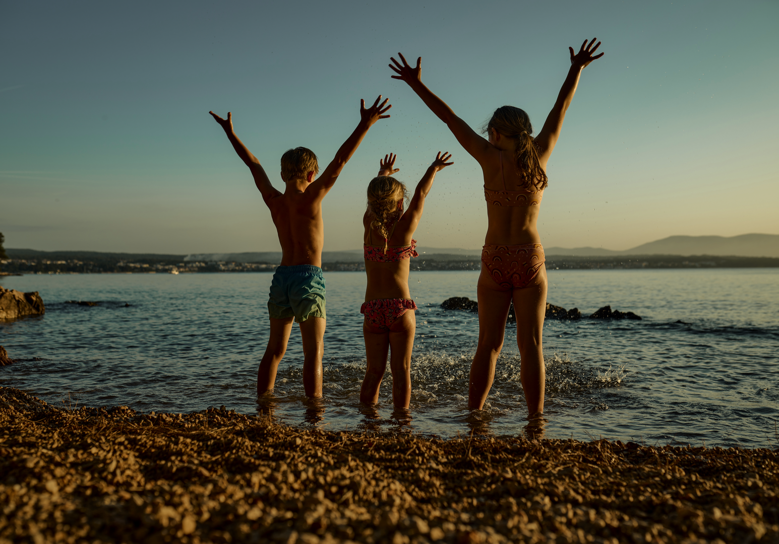 Drei Kinder stehen jubelnd am Ufer vor dem Meer.