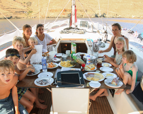 Familien und Kinder sitzen an Bord eines Segelboots und essen gemeinsam an einem  Esstisch.