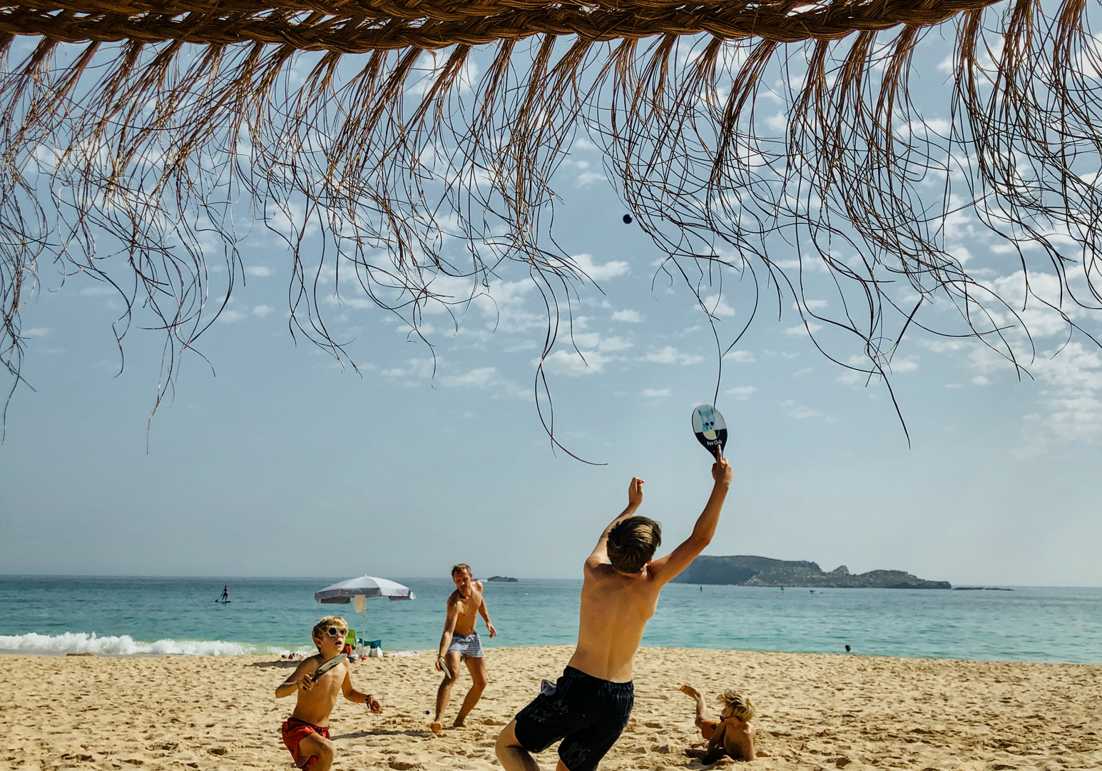 Familie spielt gemeinsam am Strand unter einem schattenspendenden Strohdach.