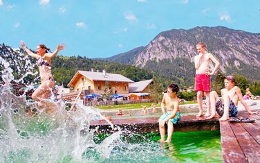 Kinder und Jugendliche haben Spaß am Naturpool. Im Hintergund sind die Unterkunft und die Berge zu sehen