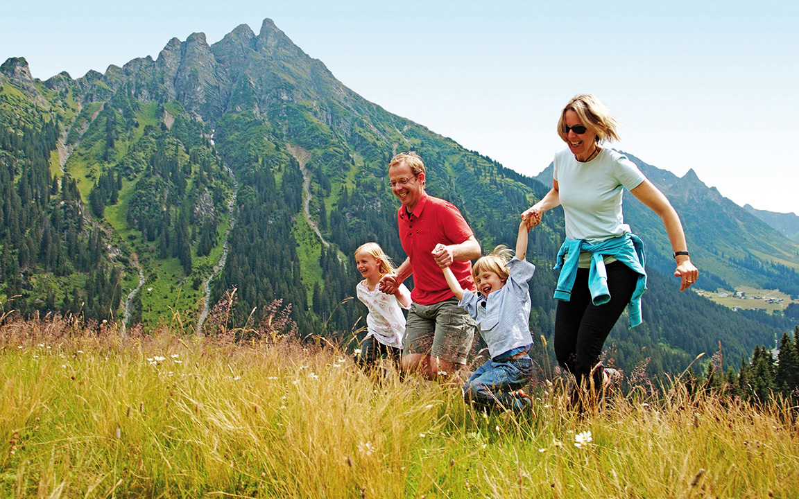 Ein Familie im Bergurlaub, die einen günstigen Urlaub gebucht hat und darum fröhlich über eine Wiese läuft.