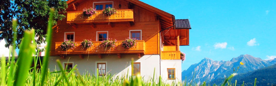 Die vamos Unterkunft wird ist umgeben von einer grünen Wiese und schönen Bergen. Die Unterkunft ist ein zweistöckiges Haus im österreicherischem Stil, mit Balkonen aus Holz, die mit Blumen dekoriert sind.